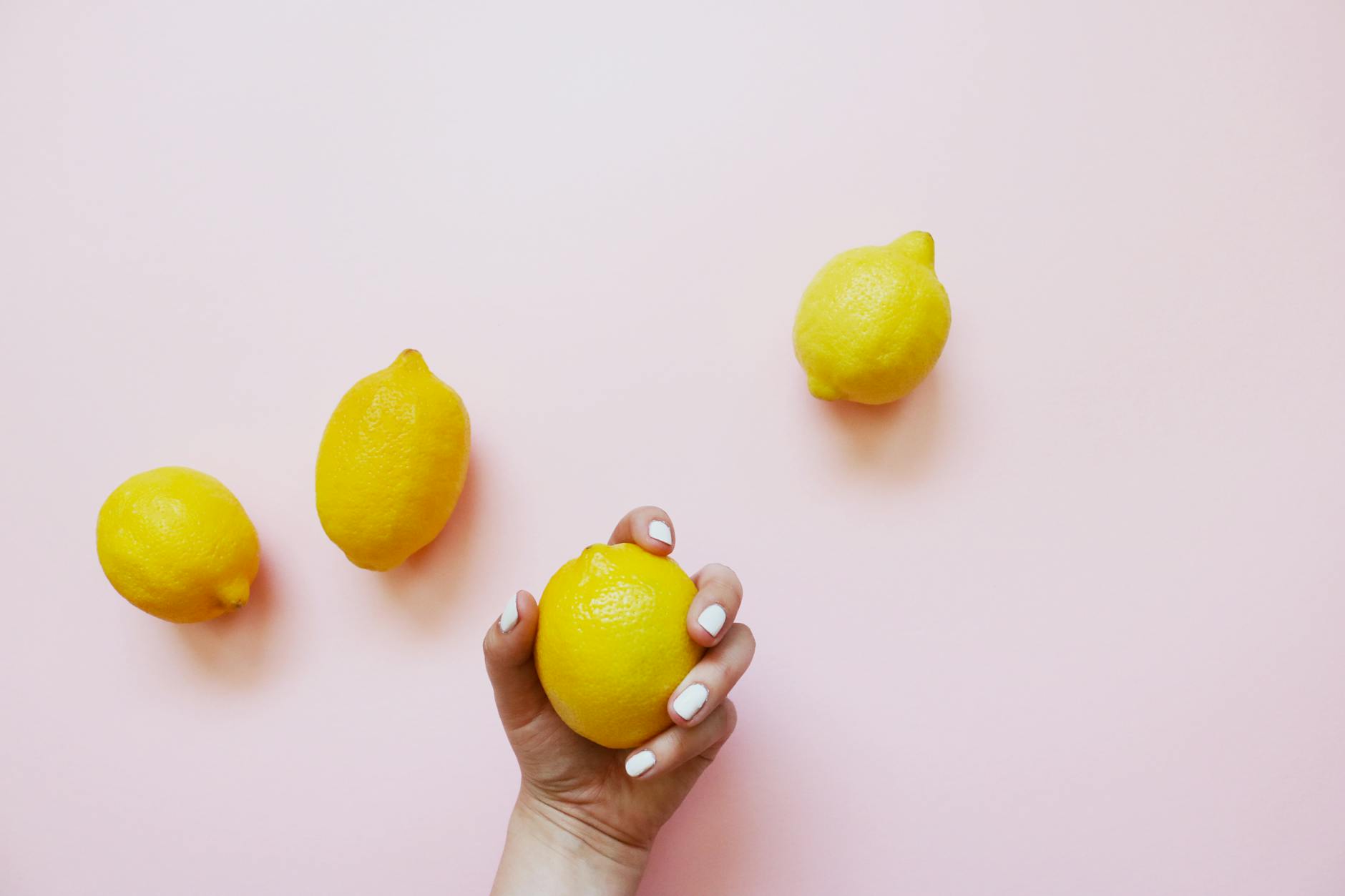 A hand with white nails holding a fresh lemon on a soft pink background.